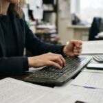 Persona escribiendo en un teclado en una oficina, con documentos, una calculadora y un teléfono sobre el escritorio, para ilustrar la gestión de cobros con software