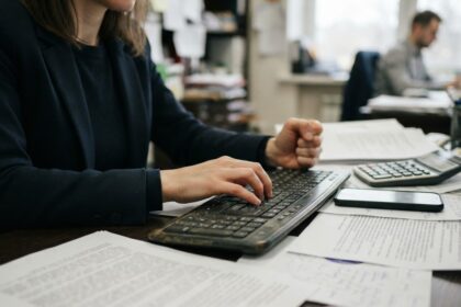 Persona escribiendo en un teclado en una oficina, con documentos, una calculadora y un teléfono sobre el escritorio, para ilustrar la gestión de cobros con software